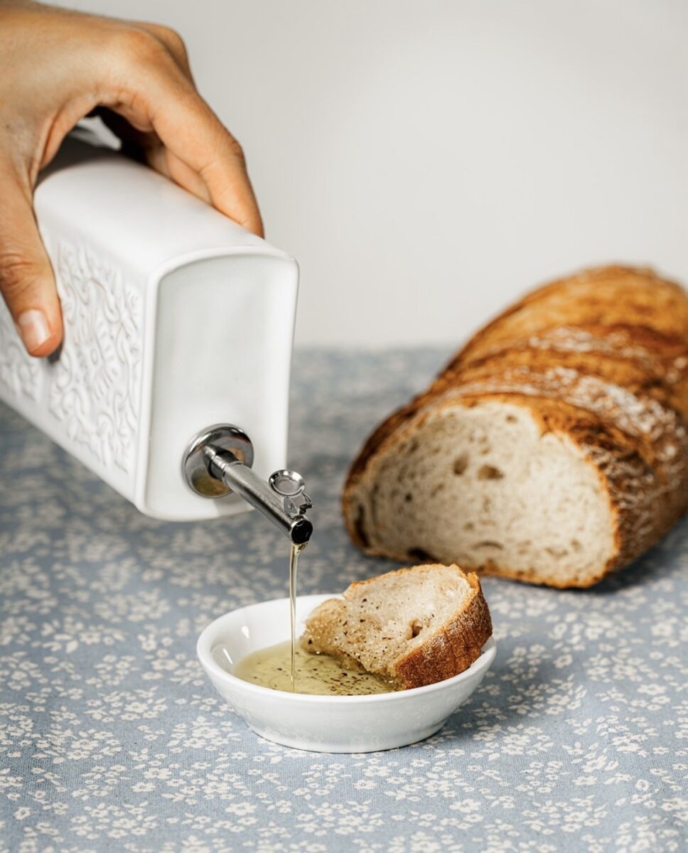 Un huilier AZULEJO blanc en céramique versant un filet d'huile d'olive sur un morceau de pain.